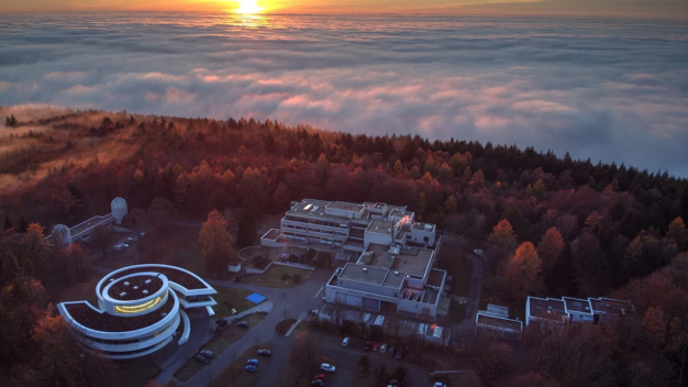 Luftbild des MPIA-Campus auf dem Königstuhl mit dem spiralförmigen Haus der Astronomie (links unten), den Teleskop-Kuppelgebäuden des Elsässer-Labors (links) und dem MPIA-Hauptgebäude (Mitte). Der Erweiterungsbau wird sich in Richtung der Kuppelgebäude an das Südende des Hauptgebäudes anschließen. (Quelle: Domink Elsässer) / Aerial view of the MPIA campus on the Königstuhl with the spiral-shaped House of Astronomy (down left), the telescope dome buildings of the Elsässer Laboratory (left) and the MPIA main building (center). The extension building will adjoin the south end of the main building in the direction of the domed buildings. (Credit: Domink Elsässer)